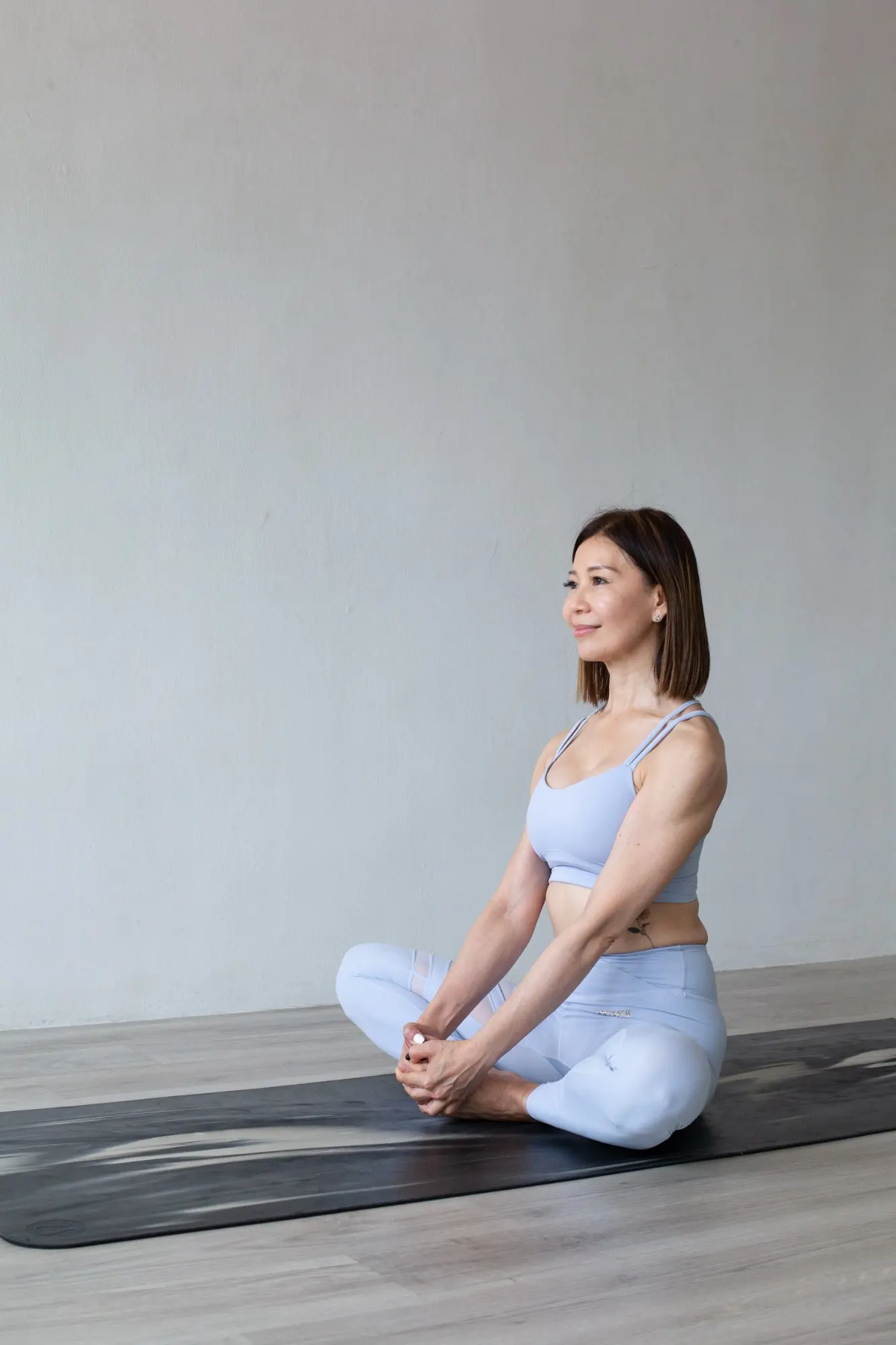 Katie sitting cross-legged on a mat in a light blue top and leggings, holding her feet